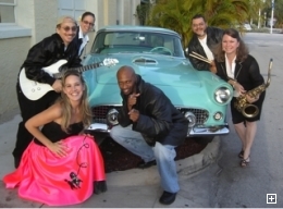 Photo of the At the Hop Oldies band dressed in 50s style clothing, posed in front of a classic 50s car